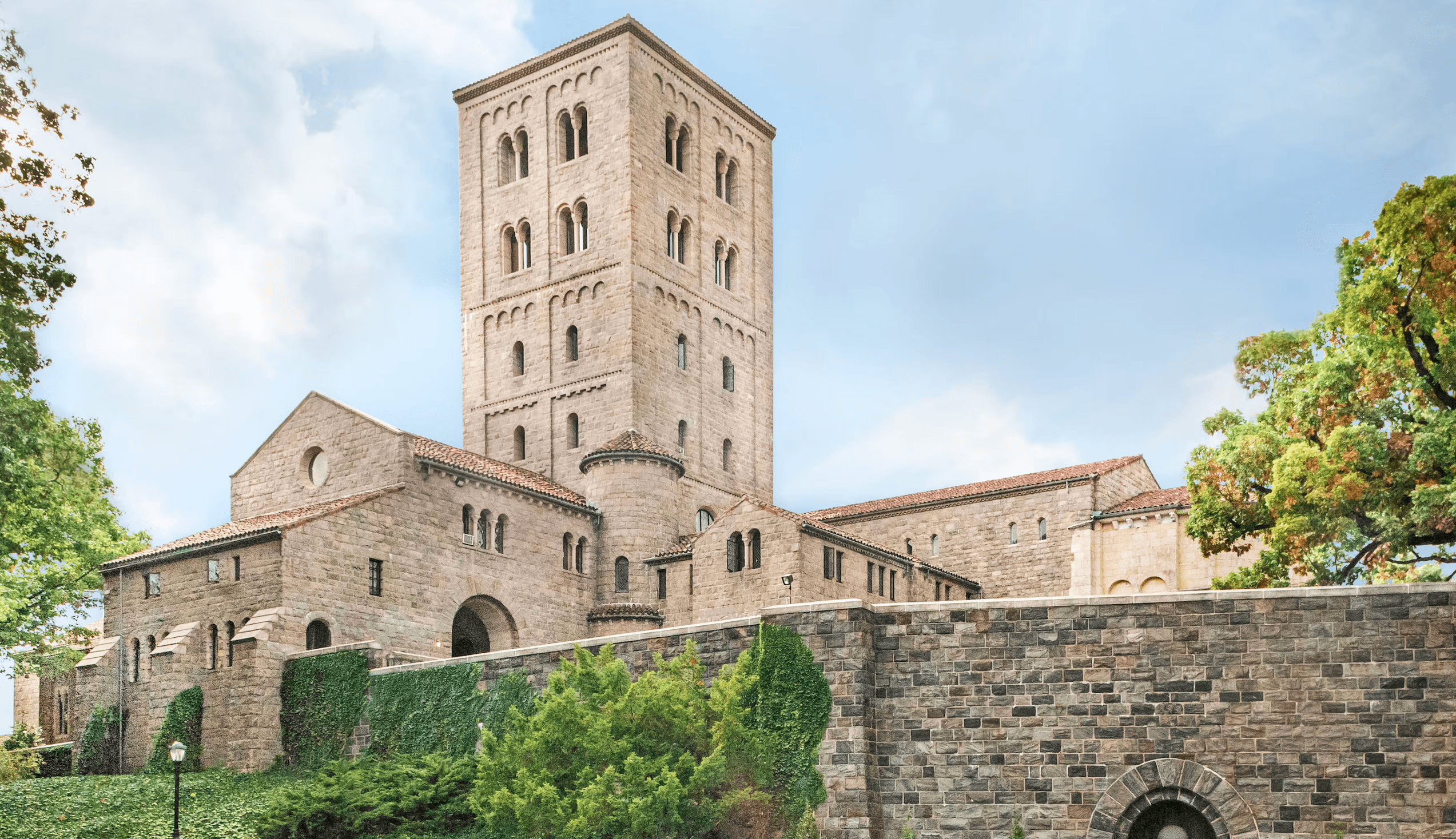 A large stone building with a tall rectangular tower and red-tiled roof, the Met Cloisters is surrounded by trees and ivy-covered stone walls under a blue sky.